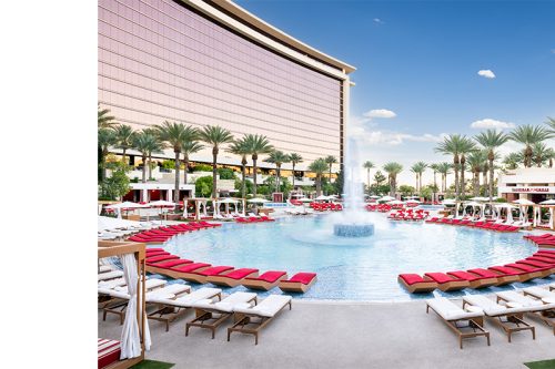 Luxury resort pool with a central fountain, palm trees, cabanas, and red lounge chairs.