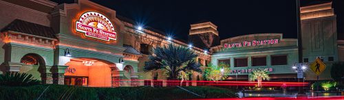 Exterior nighttime view of Santa Fe Station Hotel & Casino, illuminated with bright signage and architectural lighting, with palm trees and light trails from passing vehicles in the foreground.