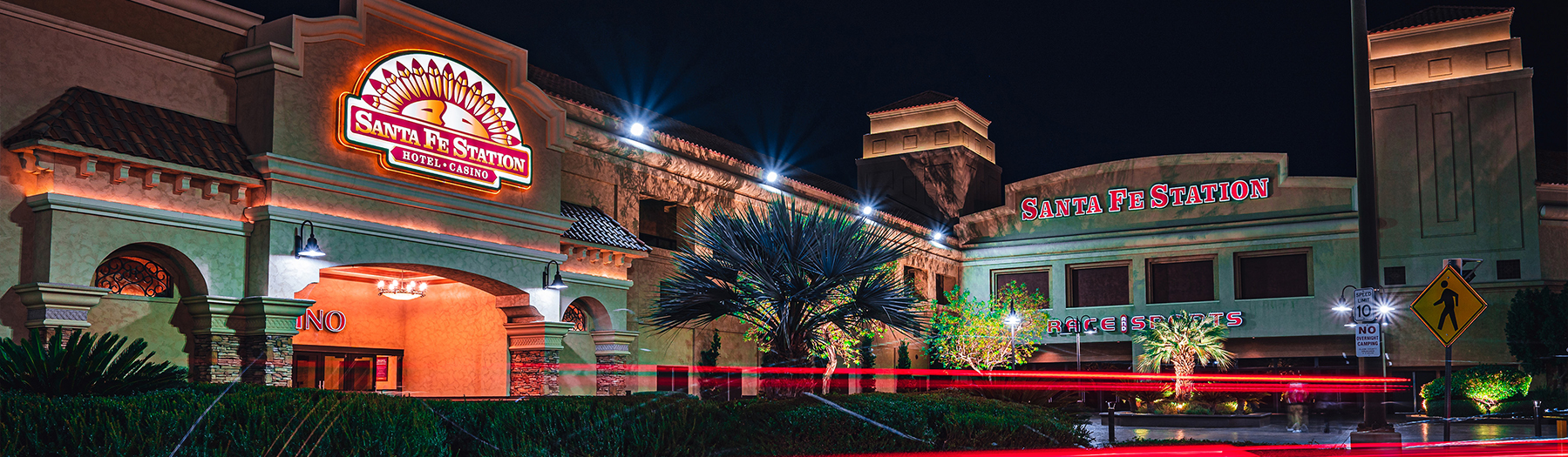 Exterior nighttime view of Santa Fe Station Hotel & Casino, illuminated with bright signage and architectural lighting, with palm trees and light trails from passing vehicles in the foreground.