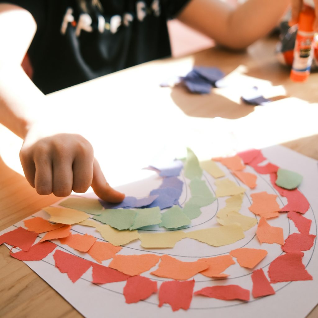 A child's hand arranges colorful torn paper pieces into a rainbow pattern on a table, creating a cheerful and creative art project.