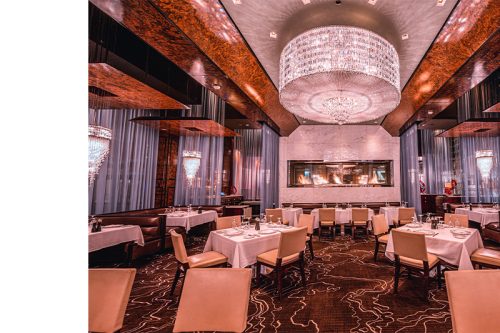 Upscale restaurant interior with white tablecloths, leather chairs, crystal chandeliers, a modern fireplace, and elegant sheer curtains.
