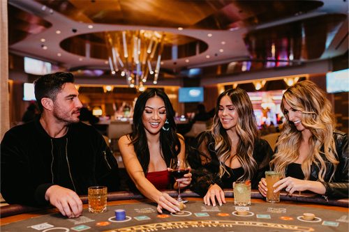 Four people seated at a casino poker table, holding drinks and engaging in the game, with a lively casino background.
