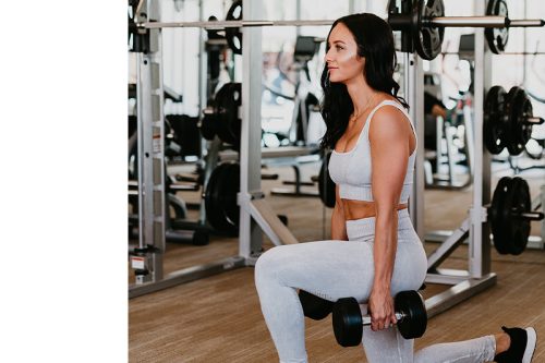 Woman in athletic wear performs a dumbbell lunge in a gym, surrounded by weightlifting equipment and racks.
