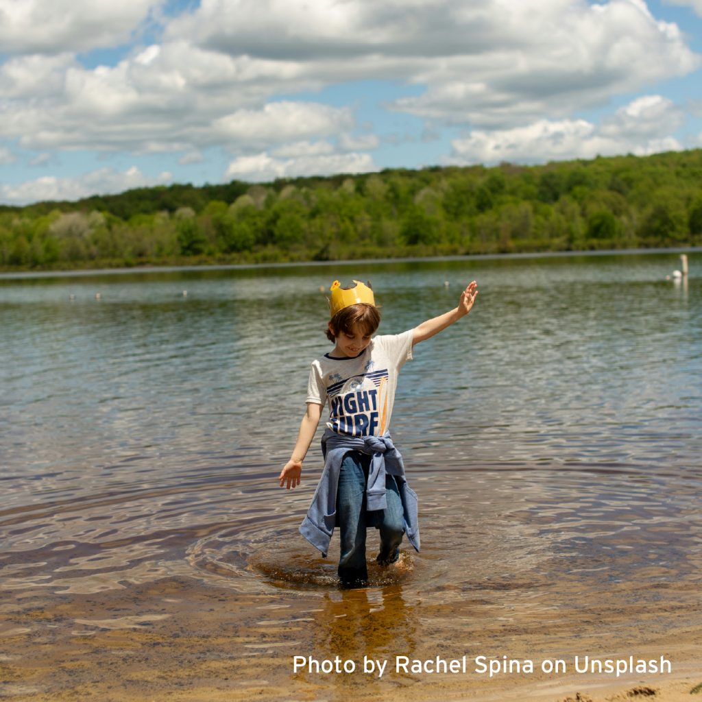 Child wearing a paper crown wades through a shallow lake on a sunny day, with trees and clouds in the background.