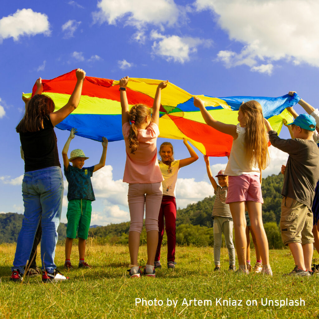 Children play together outdoors, holding a colorful parachute overhead against a bright blue sky with clouds, encouraging teamwork, movement, and fun.