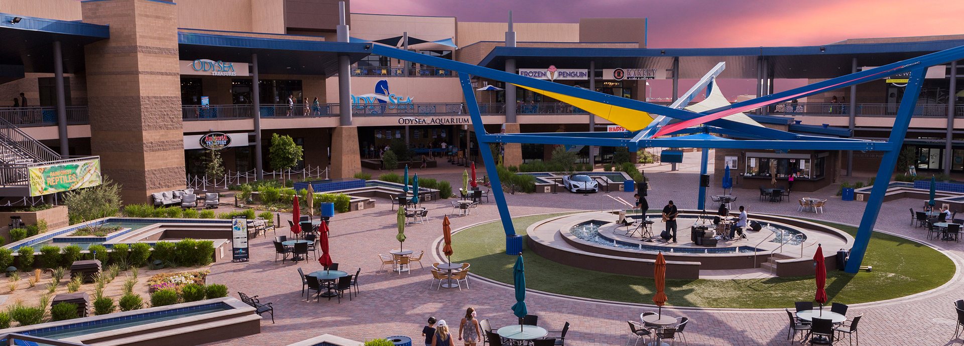 The courtyard at Arizona Boardwalk in Scottsdale at sunset, with colorful shade sails, a fountain stage and umbrella tables