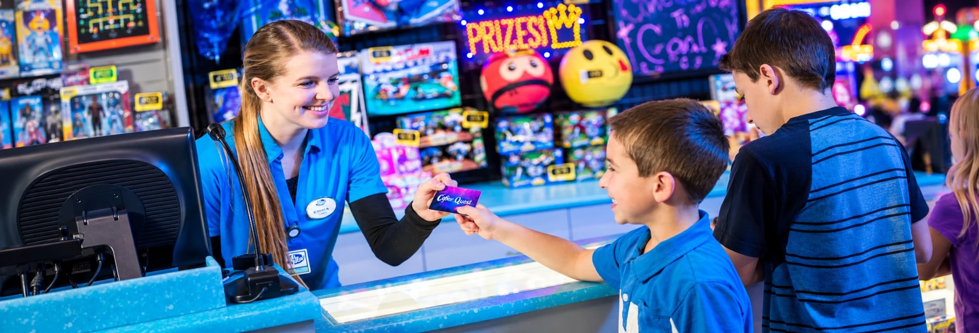 A smiling Cyber Quest teammate hands a Cyber Quest Club Card to a boy at the prize counter while his brother looks on