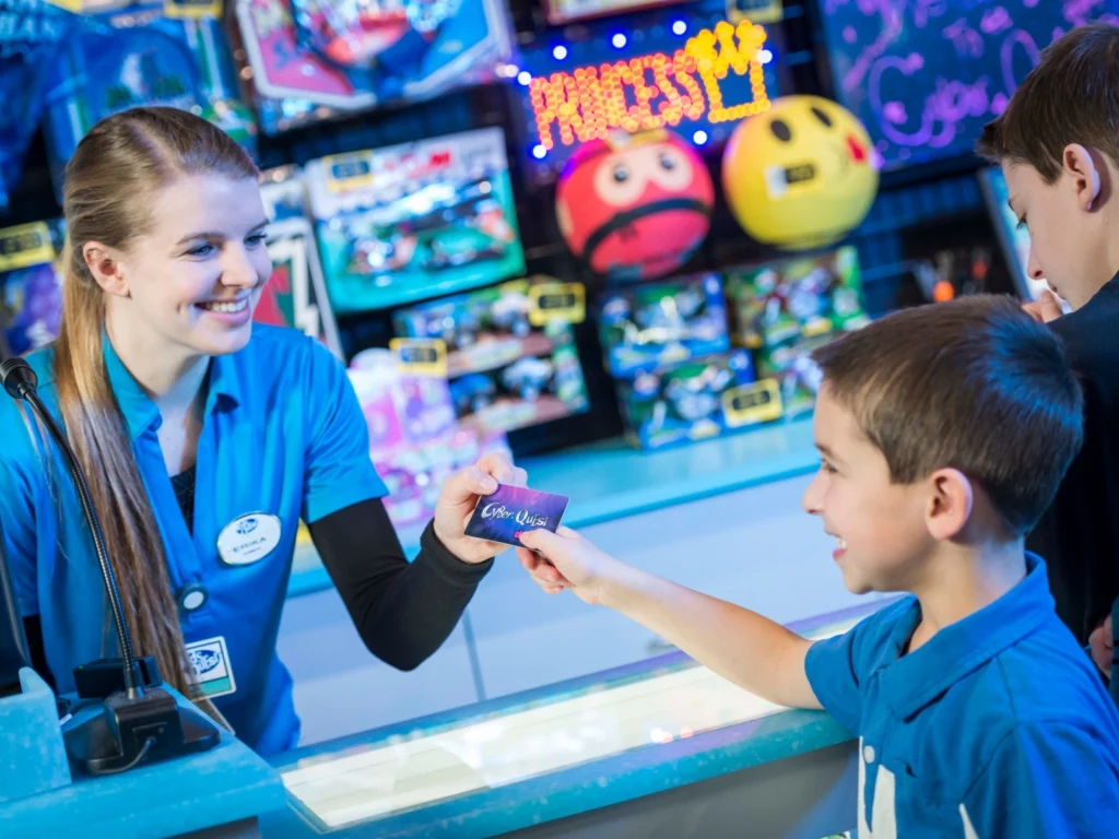 A smiling Cyber Quest teammate hands a Cyber Quest Club Card to a boy at the Prize Zone counter while his brother looks on