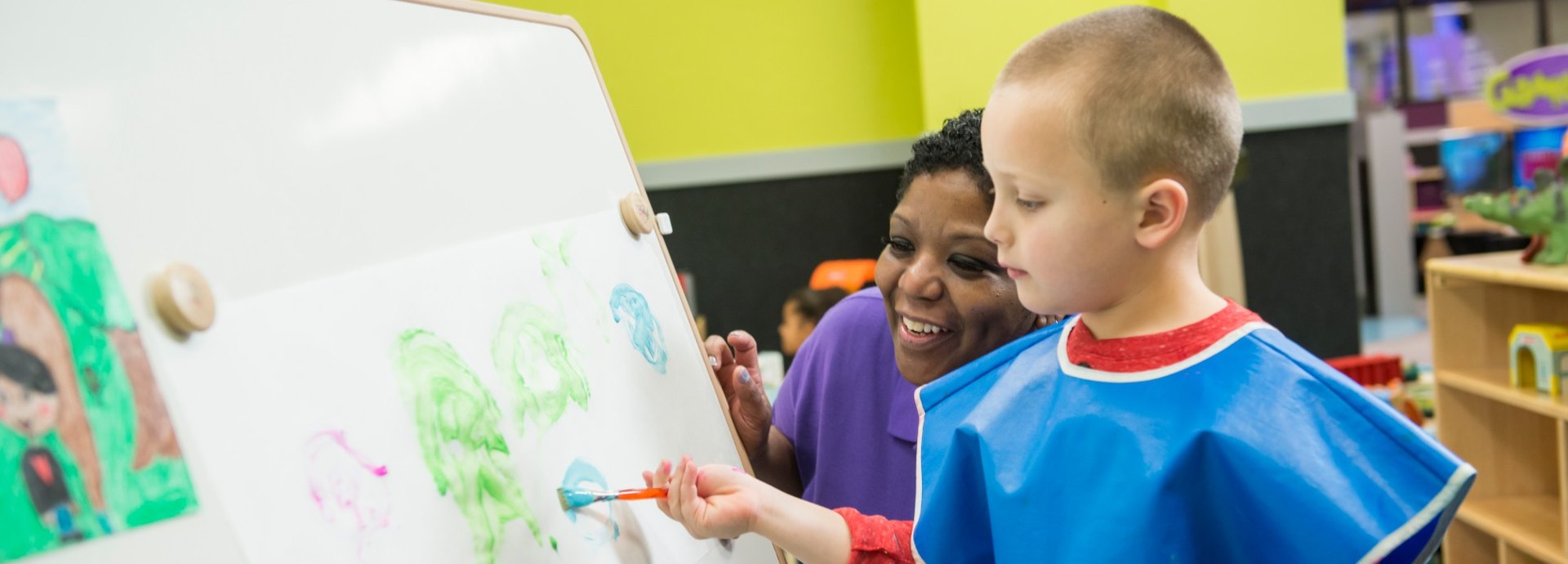 A Kids Quest teammate cheers on a boy in a paint smock painting at an easel