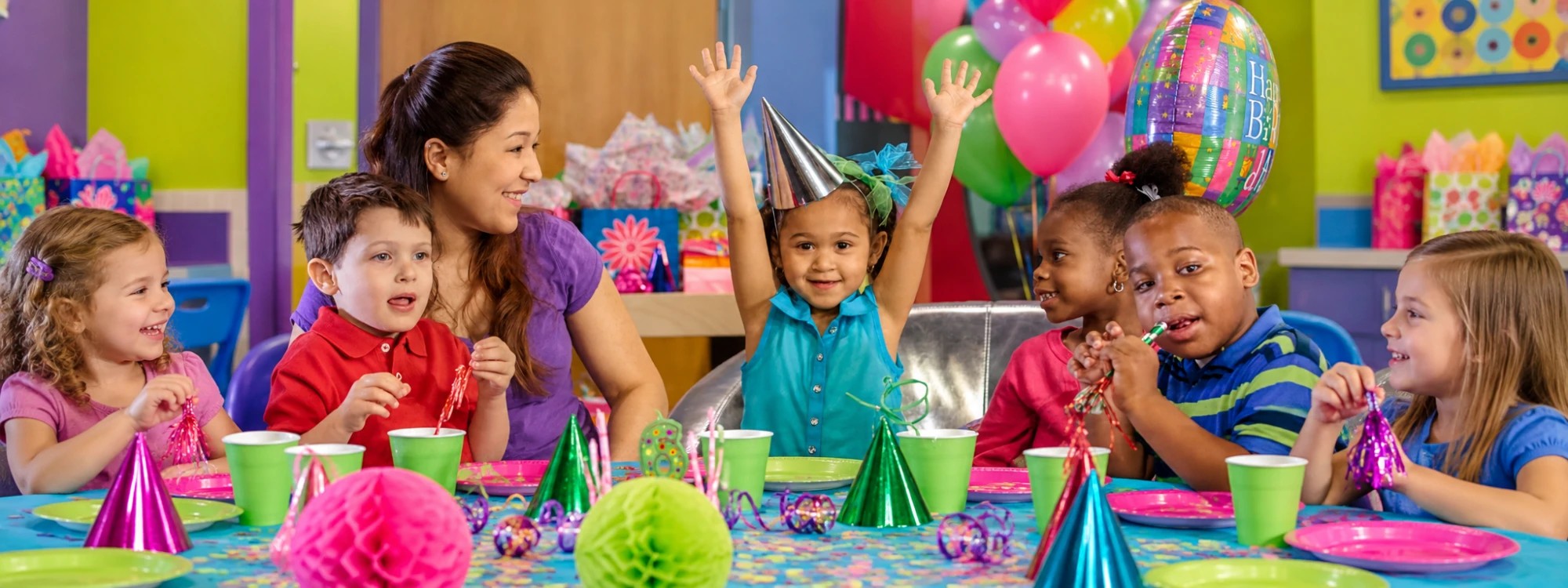 A birthday girl in a silver party hat throws her arms up at her Kids Quest party while a teammate and her friends celebrate at the party table