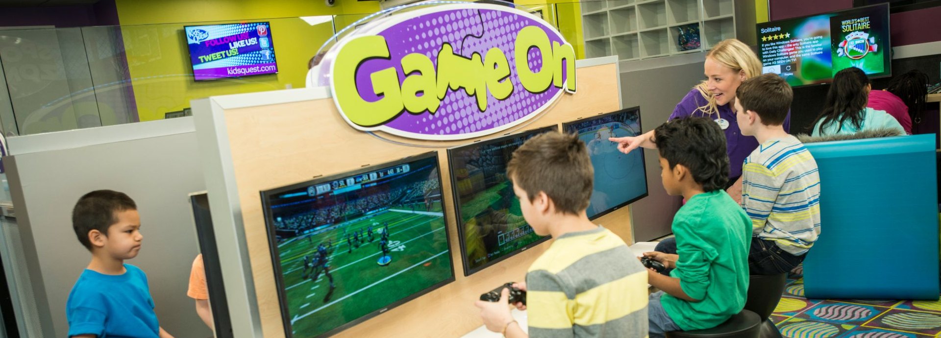 Kids play video games at the Game On station while a Kids Quest teammate points to the screen