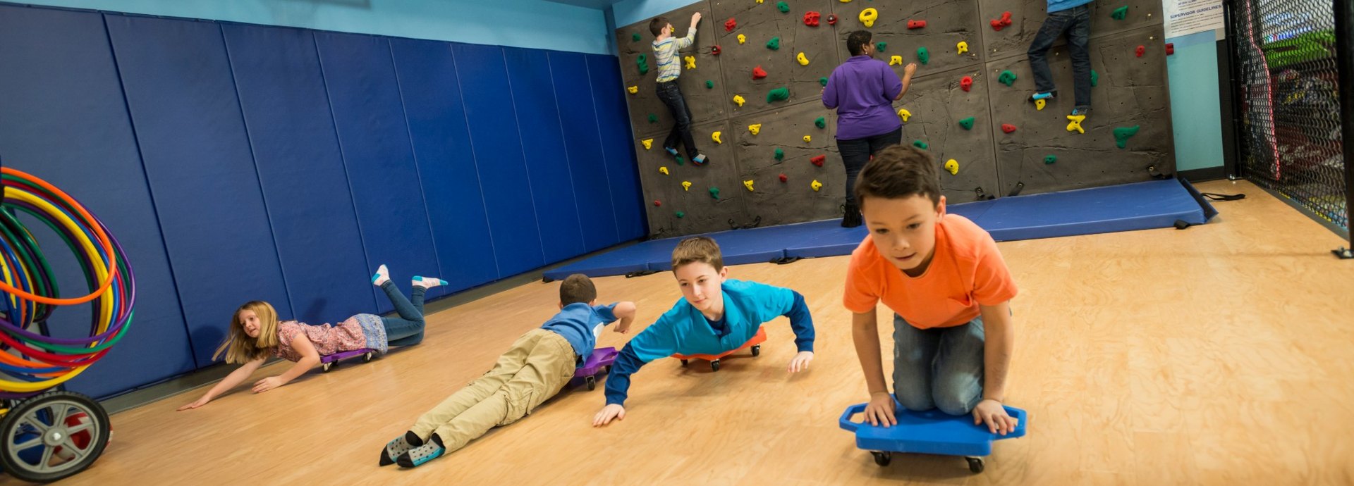 Kids racing on scooter boards across the gym at Kids Quest while others climb the rock wall with a Kids Quest teammate