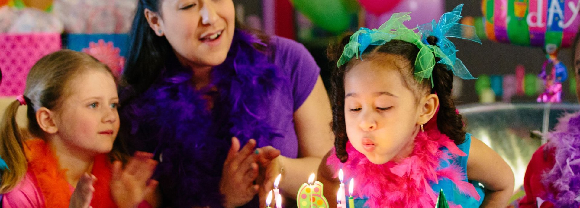 A birthday girl in a pink feather boa blows out the candles on her cake as a Kids Quest teammate and a friend clap