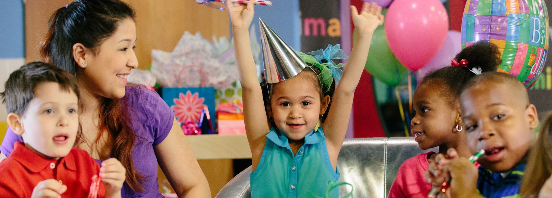 A birthday girl in a party hat throws her arms up at her Kids Quest party with friends and a teammate at the table