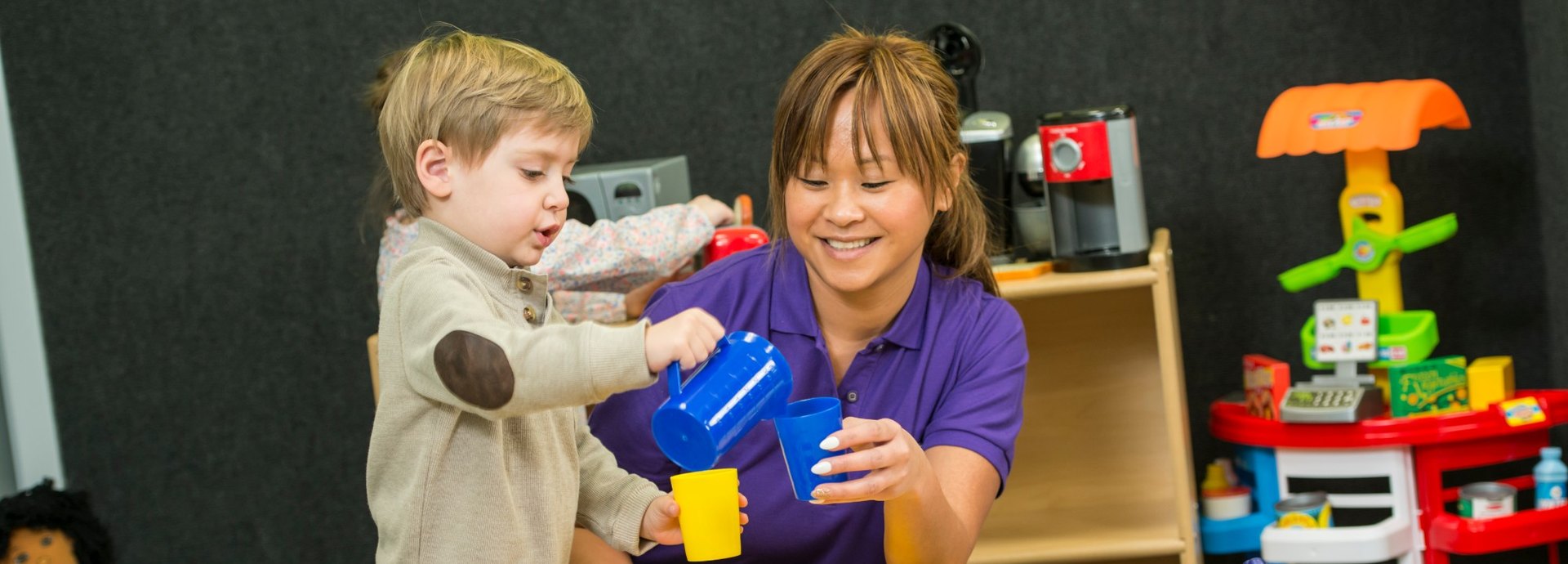 A Kids Quest teammate and a toddler pour pretend drinks at the play kitchen table