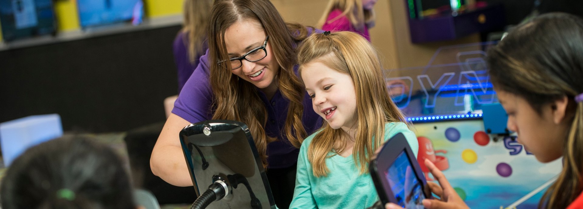A Kids Quest teammate laughs with a girl playing a tablet game while another girl plays nearby