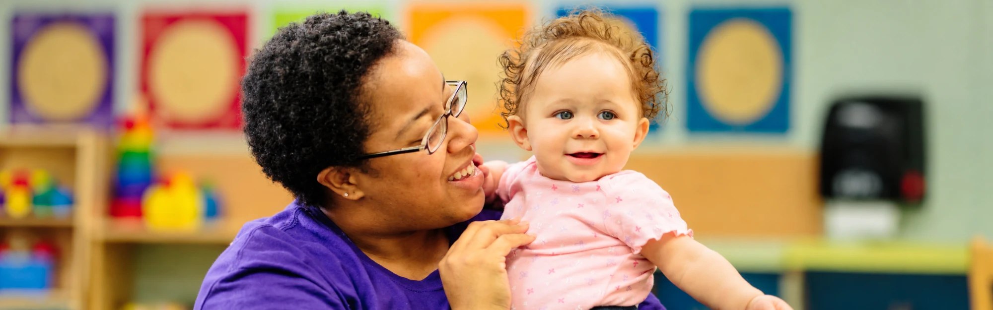 A smiling Kids Quest teammate in a purple shirt holds a curly-haired baby in the Tiny Tots room